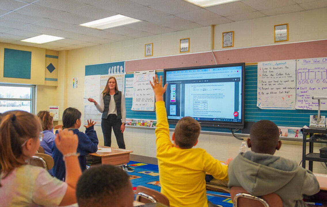 A teacher in front of her classroom with a smartboard, with a few students raising their hands