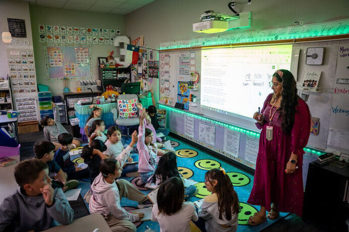 A teacher standing in front of a classroom of students sitting on a rug, some with hands raised