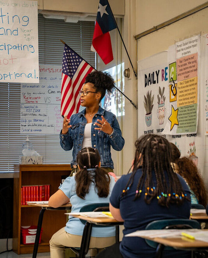 A teacher-candidate standing and speaking in front of students sitting in a row of desks