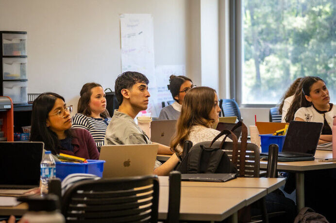 Teacher-candidates sitting at rectangular tables in an undergraduate course looking up at instructor