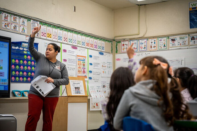 A teacher gesturing with her arm raised in front of a classroom, wh ile a few students mirror the gesture